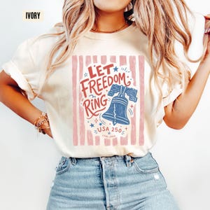 K&ouml;nnte beinhalten: Cremefarbenes T-Shirt mit patriotischem Design. Die Grafik zeigt den Text "Let Freedom Ring" mit einer blauen Freiheitsglocken-Illustration, Sternen und dem Text "USA 250 1776-2026" in Rot und Blau.