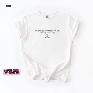 May include: A white short-sleeve t-shirt featuring the black text currently hyperfixated on hockey romance centered on the chest, accompanied by a small graphic of two crossed hockey sticks positioned directly below the printed text.