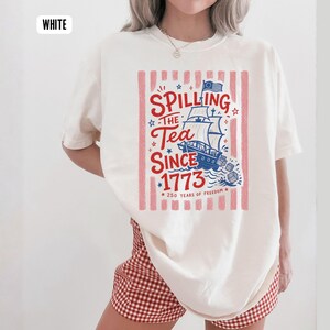 May include: White t-shirt with a red and blue graphic. The graphic features a ship, stars, and the text "Spilling the Tea Since 1773" and "250 Years of Freedom". The shirt has a crew neck and short sleeves.