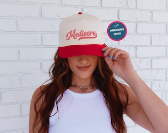 May include: A cream and red baseball cap with the word "Mediocre" in a cursive font. The cap has a red brim and a red button on top. The image also includes a small, round sticker that says "Embroidered Design".