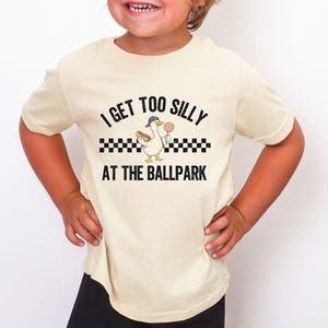 May include: A cream-colored t-shirt with the text "I GET TOO SILLY AT THE BALLPARK". The graphic features a cartoon duck wearing a baseball cap, holding a hot dog and a lollipop. The design includes a black and white checkered pattern.