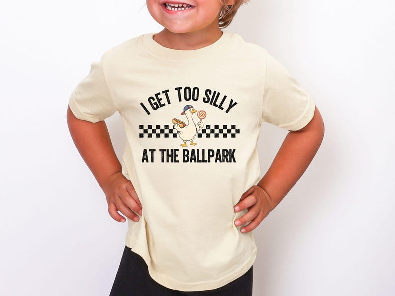 May include: A cream-colored t-shirt with the text "I GET TOO SILLY AT THE BALLPARK". The graphic features a cartoon duck wearing a baseball cap, holding a hot dog and a lollipop. The design includes a black and white checkered pattern.