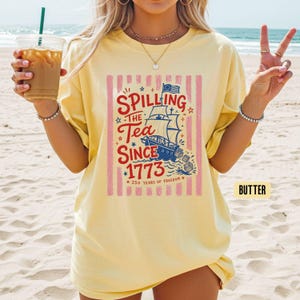 May include: Butter-colored t-shirt with a graphic that reads "Spilling the Tea Since 1773" featuring a ship, stars, and stripes. The shirt has a relaxed fit and is worn on a beach.