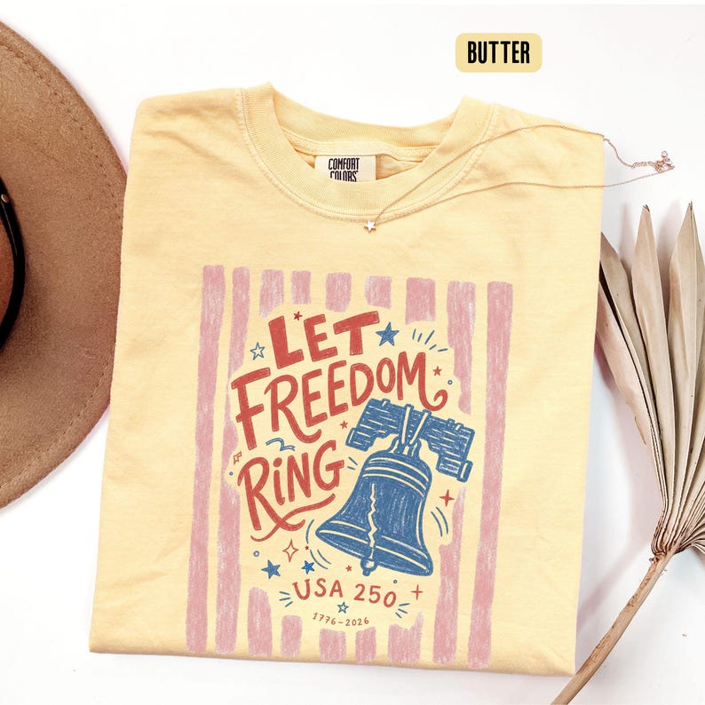 K&ouml;nnte beinhalten: Hellgelbes T-Shirt mit patriotischem Design. Die Grafik zeigt die Worte "Let Freedom Ring" in Rot und Blau, eine blaue Freiheitsglocke und den Text "USA 250" und "1776-2026". Eine goldene Halskette mit Sternanh&auml;nger ist ebenfalls zu sehen.