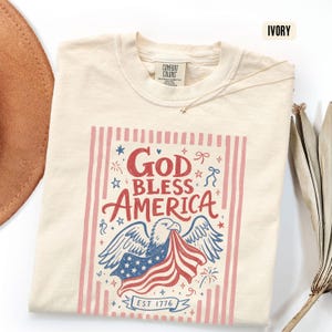 May include: Ivory-colored t-shirt with a patriotic design. The graphic features an eagle holding a flag with the text "God Bless America" and "EST 1776" in red and blue. The shirt has a crew neck and short sleeves.