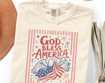 May include: Ivory-colored t-shirt with a patriotic design. The graphic features an eagle holding a flag with the text "God Bless America" and "EST 1776" in red and blue. The shirt has a crew neck and short sleeves.