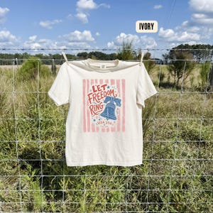 K&ouml;nnte beinhalten: Elfenbeinfarbenes T-Shirt mit dem Text "Let Freedom Ring" in Rot und Blau, mit einer Glockengrafik und vertikalen roten Streifen. Das T-Shirt h&auml;ngt drau&szlig;en an einer W&auml;scheleine.