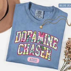 Pode incluir: Uma camiseta azul jeans Comfort Colors com as palavras "Dopamine Chaser" em um design floral. A camisa tamb&eacute;m tem a palavra "ADHD" e um colar de estrela dourada. Um chap&eacute;u marrom &eacute; vis&iacute;vel no fundo.