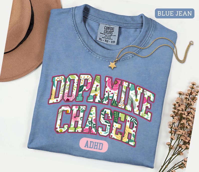 Pode incluir: Uma camiseta azul jeans Comfort Colors com as palavras "Dopamine Chaser" em um design floral. A camisa tamb&eacute;m tem a palavra "ADHD" e um colar de estrela dourada. Um chap&eacute;u marrom &eacute; vis&iacute;vel no fundo.