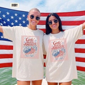 May include: Ivory-colored t-shirts with the text "God Bless America" and an eagle graphic. The design incorporates red, white, and blue colors, with the text "EST 1776". The shirts are worn in front of an American flag.