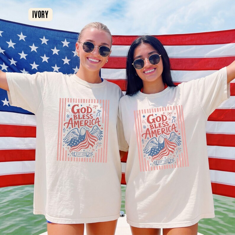 May include: Ivory-colored t-shirts with the text "God Bless America" and an eagle graphic. The design incorporates red, white, and blue colors, with the text "EST 1776". The shirts are worn in front of an American flag.