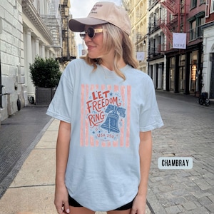 K&ouml;nnte beinhalten: Hellblaues T-Shirt mit dem Text "Let Freedom Ring" in Rot und Blau, mit einer Glockengrafik. Das Shirt hat vertikale rot-wei&szlig;e Streifen und den Text "USA 250".