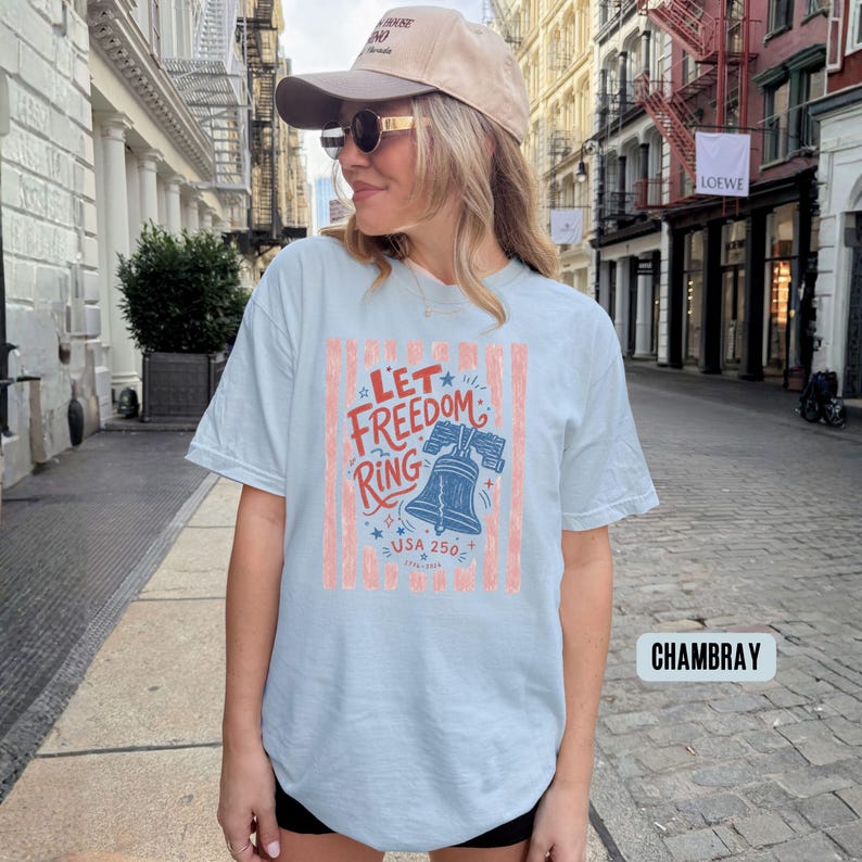 K&ouml;nnte beinhalten: Hellblaues T-Shirt mit dem Text "Let Freedom Ring" in Rot und Blau, mit einer Glockengrafik. Das Shirt hat vertikale rot-wei&szlig;e Streifen und den Text "USA 250".