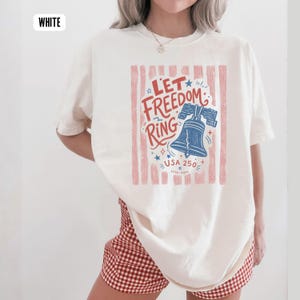 K&ouml;nnte beinhalten: Cremefarbenes T-Shirt mit patriotischem Design. Die Grafik zeigt den Text "Let Freedom Ring" mit einer blauen Liberty Bell, Sternen und dem Text "USA 250" in Rot und Blau. Das Shirt hat eine lockere Passform.