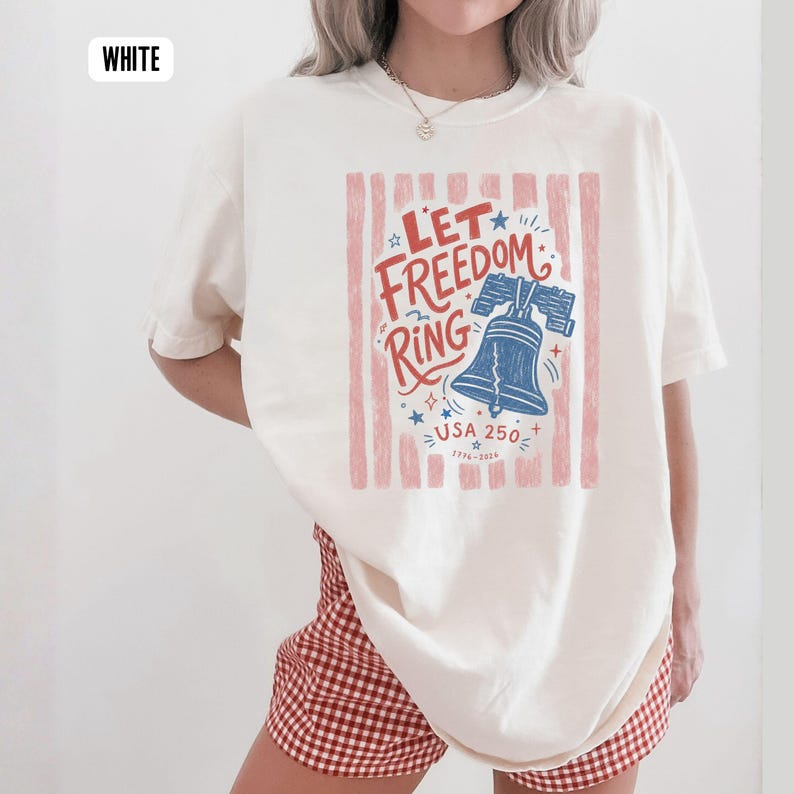 K&ouml;nnte beinhalten: Cremefarbenes T-Shirt mit patriotischem Design. Die Grafik zeigt den Text "Let Freedom Ring" mit einer blauen Liberty Bell, Sternen und dem Text "USA 250" in Rot und Blau. Das Shirt hat eine lockere Passform.
