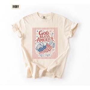May include: Ivory-colored t-shirt with a patriotic design. The graphic features an eagle holding a waving American flag, with the words "God Bless America" in red and blue. The shirt includes the text "EST 1776".