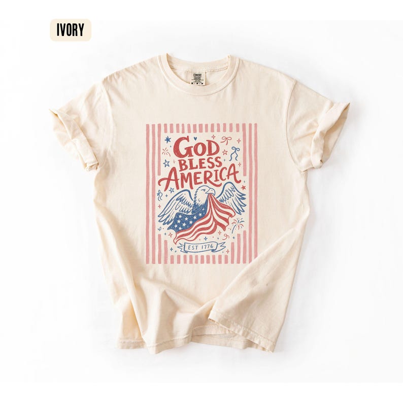 May include: Ivory-colored t-shirt with a patriotic design. The graphic features an eagle holding a waving American flag, with the words "God Bless America" in red and blue. The shirt includes the text "EST 1776".