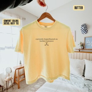 May include: A butter yellow short-sleeve t-shirt with a crew neckline. The center of the chest features the black text 'currently hyperfixated on hockey romance' above a small graphic of two crossed hockey sticks. The shirt is displayed on a hanger.