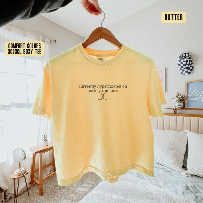 May include: A butter yellow short-sleeve t-shirt with a crew neckline. The center of the chest features the black text 'currently hyperfixated on hockey romance' above a small graphic of two crossed hockey sticks. The shirt is displayed on a hanger.
