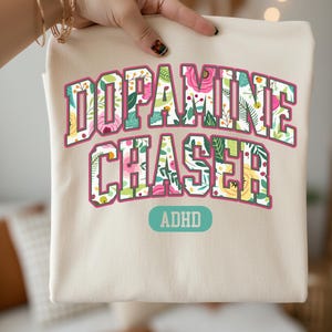 Pode incluir: Camiseta cor marfim com as palavras "Dopamine Chaser" em um design floral, com a palavra "ADHD" abaixo. O texto est&aacute; em uma fonte de estilo universit&aacute;rio com uma borda rosa.
