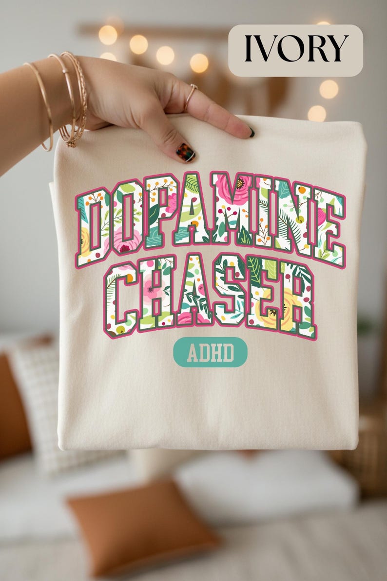 Pode incluir: Camiseta cor marfim com as palavras "Dopamine Chaser" em um design floral, com a palavra "ADHD" abaixo. O texto est&aacute; em uma fonte de estilo universit&aacute;rio com uma borda rosa.