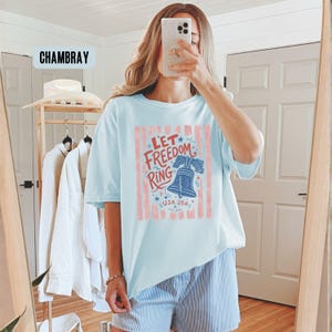 K&ouml;nnte beinhalten: Hellblaues T-Shirt mit dem Text "Let Freedom Ring" in Rot und Blau, mit einer Glockengrafik. Das Shirt hat ein USA 250 Logo und rote vertikale Streifen.