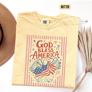 May include: Butter-colored t-shirt with a patriotic design. The graphic features an eagle holding an American flag, the text "God Bless America," and the year "1776." A gold necklace with a star pendant is draped on the shirt.