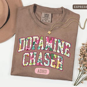 Pode incluir: Camiseta cor de caf&eacute; com as palavras "Dopamine Chaser" em um design floral. A camisa tamb&eacute;m tem a palavra "ADHD". Um colar de ouro com um pingente de estrela e um chap&eacute;u marrom tamb&eacute;m est&atilde;o na imagem.