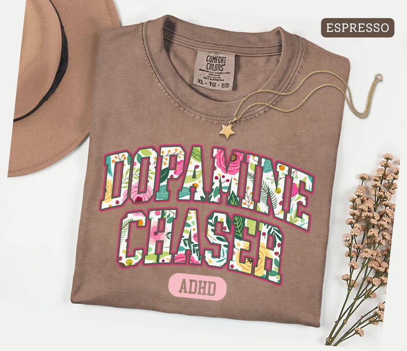 Pode incluir: Camiseta cor de caf&eacute; com as palavras "Dopamine Chaser" em um design floral. A camisa tamb&eacute;m tem a palavra "ADHD". Um colar de ouro com um pingente de estrela e um chap&eacute;u marrom tamb&eacute;m est&atilde;o na imagem.