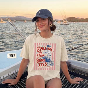 May include: Ivory-colored t-shirt with a graphic that reads "Spilling the Tea Since 1773" featuring a ship and stars in red, white, and blue. The shirt is worn by a person on a boat, with a blue cap and gold earrings.
