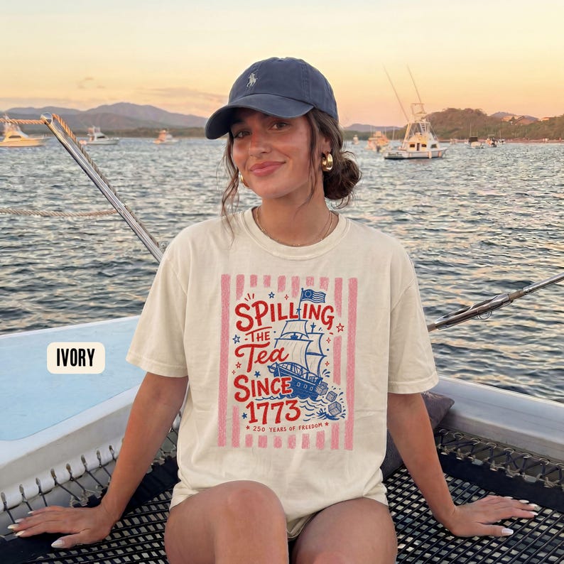 May include: Ivory-colored t-shirt with a graphic that reads "Spilling the Tea Since 1773" featuring a ship and stars in red, white, and blue. The shirt is worn by a person on a boat, with a blue cap and gold earrings.