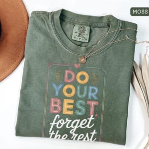 May include: A moss green t-shirt with the words "DO YOUR BEST forget the rest" in a colorful, playful font. The shirt has a crew neck and short sleeves. A gold necklace with a pendant is draped across the shirt.
