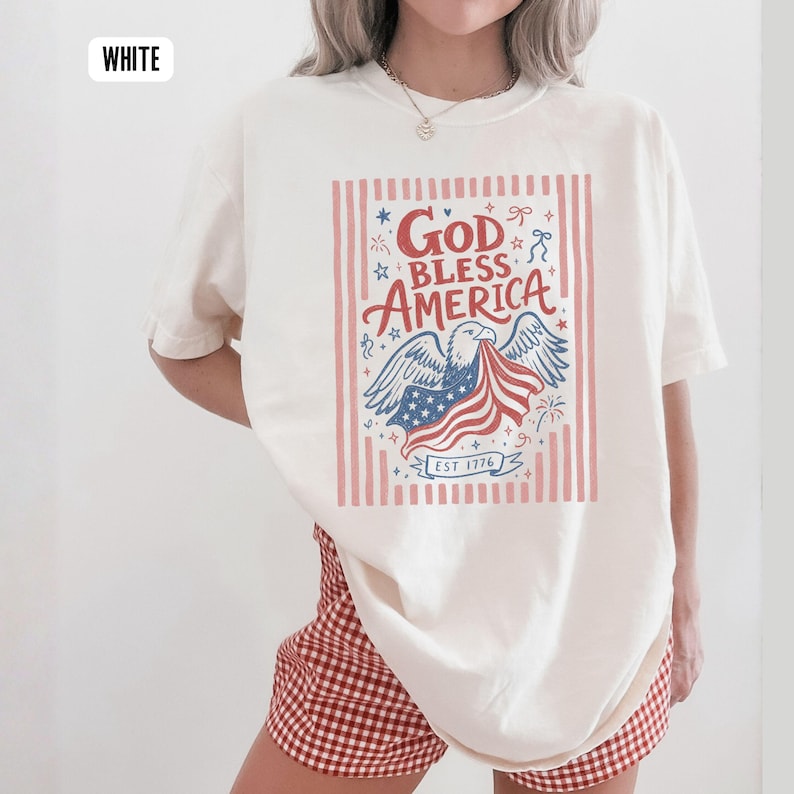 May include: White t-shirt with a patriotic design. The graphic features an eagle holding a waving American flag, with the words "God Bless America" and "EST 1776". The design is framed by red and white stripes.