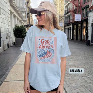 May include: Light blue t-shirt with a graphic that says "God Bless America" with an eagle and American flag design. The shirt has a classic crew neck and short sleeves. The text "EST 1776" is also included in the design.