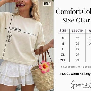 May include: A woman wearing an ivory-colored boxy tee shirt and white skirt, holding a straw tote bag with a pink flower. The image includes a size chart with measurements in inches for the tee shirt, ranging from S to 2XL.