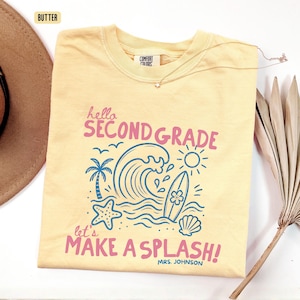 May include: A butter yellow short-sleeve t-shirt featuring a graphic with the text "hello SECOND GRADE" and "let's MAKE A SPLASH! MRS. JOHNSON". The design includes a blue wave, palm tree, surfboard, starfish, seashell, and sun.