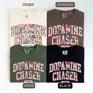 Pode incluir: Quatro camisetas nas cores marfim, caf&eacute;, musgo e preto. Cada camisa apresenta o texto "DOPAMINE CHASER" em um design floral. Gola redonda.