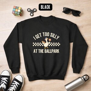 May include: Black sweatshirt with the text "I Get Too Silly At The Ballpark" and a cartoon duck wearing a hat and holding a lollipop. The sweatshirt has long sleeves and a crew neck. The word "BLACK" is in a black rectangle at the top.
