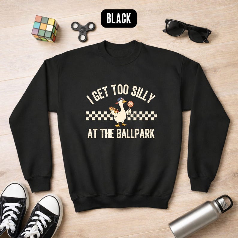 May include: Black sweatshirt with the text "I Get Too Silly At The Ballpark" and a cartoon duck wearing a hat and holding a lollipop. The sweatshirt has long sleeves and a crew neck. The word "BLACK" is in a black rectangle at the top.