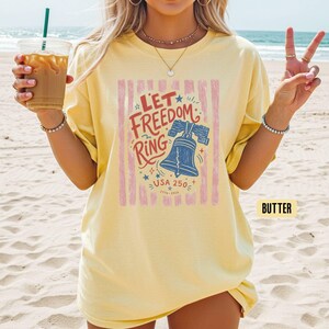 K&ouml;nnte beinhalten: Hellgelbes T-Shirt mit patriotischem Design. Die Grafik zeigt eine blaue Liberty Bell, den Text "Let Freedom Ring" und "USA 250" mit den Jahren 1776-2026. Das Design ist vor rosa vertikalen Streifen.
