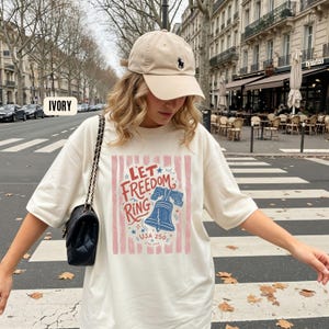 K&ouml;nnte beinhalten: Cremefarbenes T-Shirt mit dem Schriftzug "Let Freedom Ring" und einer Grafik der Liberty Bell. Das Shirt wird mit einer beigefarbenen Baseballkappe mit schwarzem Logo getragen. Eine schwarze Handtasche mit Kettenriemen ist ebenfalls zu sehen.