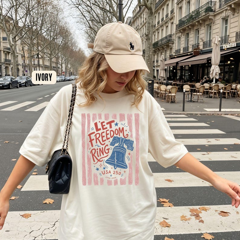 K&ouml;nnte beinhalten: Cremefarbenes T-Shirt mit dem Schriftzug "Let Freedom Ring" und einer Grafik der Liberty Bell. Das Shirt wird mit einer beigefarbenen Baseballkappe mit schwarzem Logo getragen. Eine schwarze Handtasche mit Kettenriemen ist ebenfalls zu sehen.