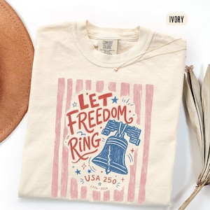 K&ouml;nnte beinhalten: Cremefarbenes T-Shirt mit dem Text "Let Freedom Ring" in Rot und Blau, mit einer Grafik der Freiheitsglocke. Das Design umfasst rosa vertikale Streifen und den Text "USA 250+ 1776-2026".