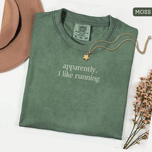 Może przedstawiać: Zielona koszulka w kolorze mchu z białym napisem "apparently, i like running". Złoty naszyjnik z gwiazdką i brązowy kapelusz leżą obok koszulki. Koszulka w rozmiarze XL-TG-EG.