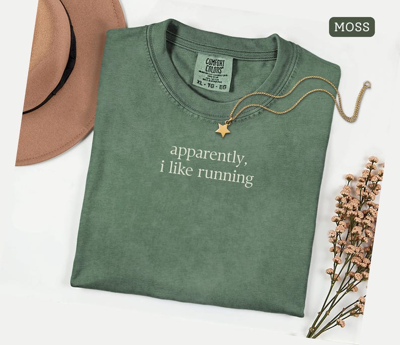 Może przedstawiać: Zielona koszulka w kolorze mchu z białym napisem "apparently, i like running". Złoty naszyjnik z gwiazdką i brązowy kapelusz leżą obok koszulki. Koszulka w rozmiarze XL-TG-EG.