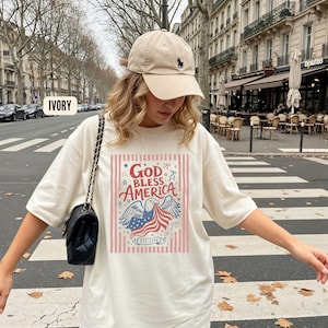 May include: Ivory-colored t-shirt with the text "GOD BLESS AMERICA" in red and blue, featuring an eagle and American flag design. The shirt has a crew neck and short sleeves. A beige baseball cap with a black logo is also visible.