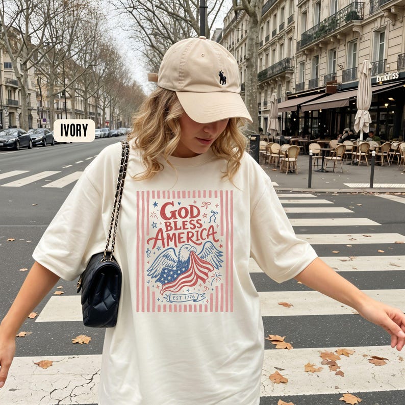 May include: Ivory-colored t-shirt with the text "GOD BLESS AMERICA" in red and blue, featuring an eagle and American flag design. The shirt has a crew neck and short sleeves. A beige baseball cap with a black logo is also visible.