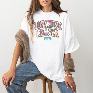 Pode incluir: Uma camiseta branca com o texto "DOPAMINE CHASER" em um design floral, com "ADHD" abaixo. A camiseta &eacute; usada com jeans azuis e um su&eacute;ter marrom sobre os ombros.