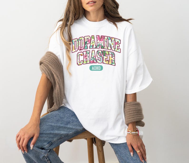 Pode incluir: Uma camiseta branca com o texto "DOPAMINE CHASER" em um design floral, com "ADHD" abaixo. A camiseta &eacute; usada com jeans azuis e um su&eacute;ter marrom sobre os ombros.