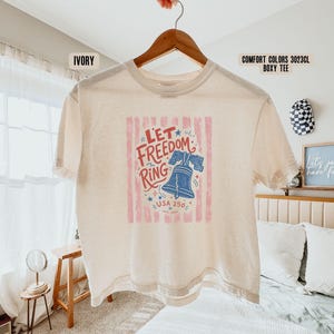 K&ouml;nnte beinhalten: Elfenbeinfarbenes Comfort Colors Boxy-T-Shirt mit der Aufschrift "Let Freedom Ring" und einer blauen Liberty Bell-Illustration. Die Grafik enth&auml;lt auch den Text "USA 250" und ist von rot-wei&szlig;en Streifen umrahmt.